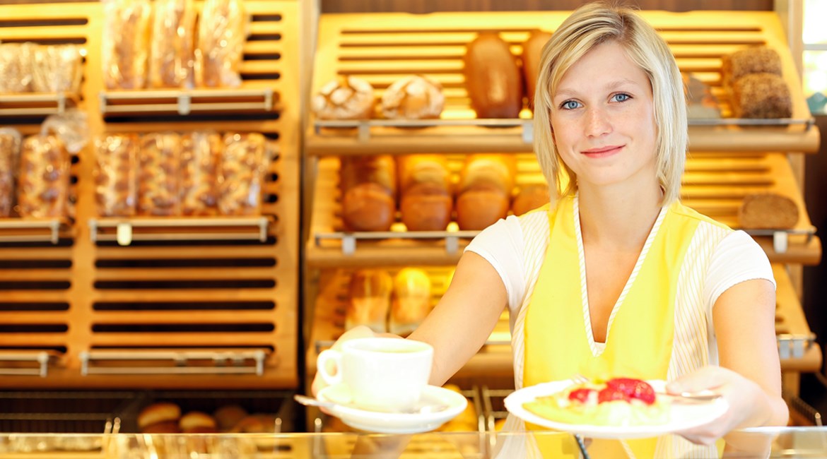 Image of woman in yellow apron smiling and offering baked goods from behind the counter of the business