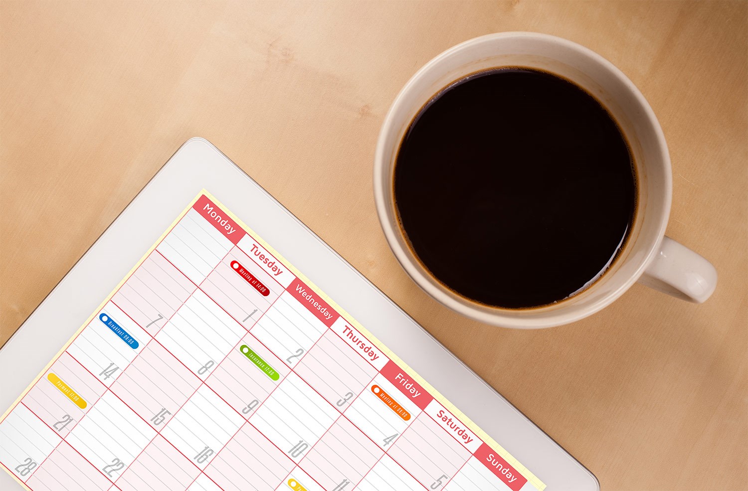 Image of coffee cup filled with coffee sitting on desk next to calendar with days of the month and different colors coded on each day.